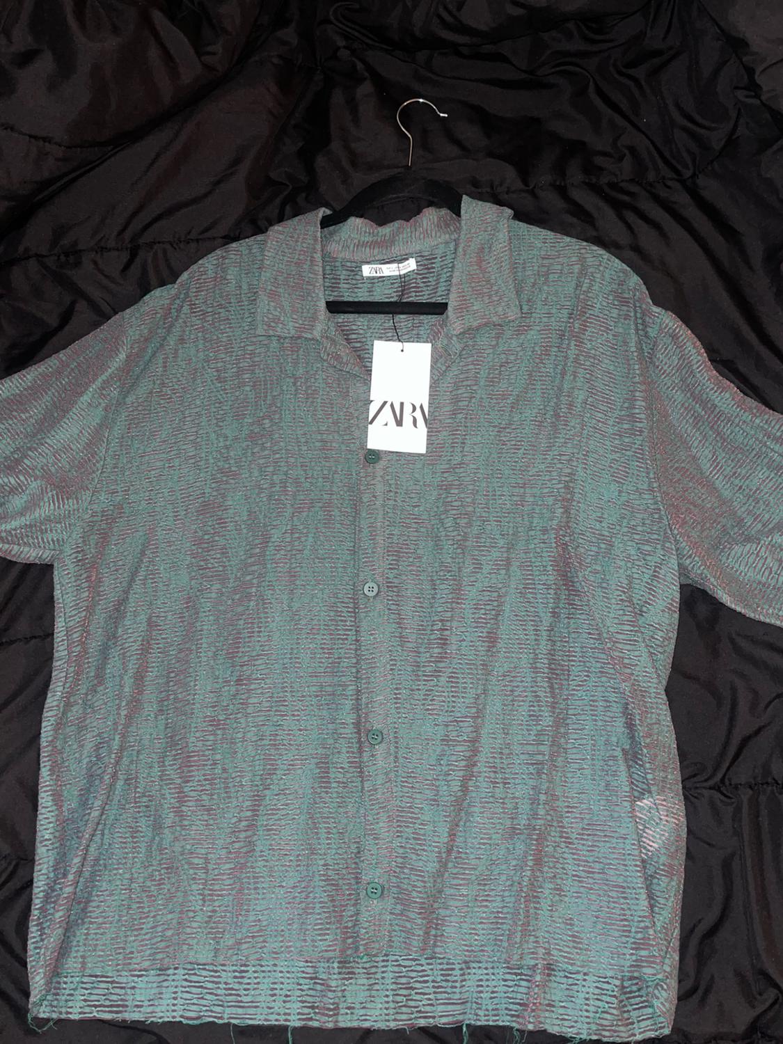 Textured green short-sleeve shirt with buttons, hanging on a hanger over wrinkled black fabric. Includes a tag with the text ZARA.
