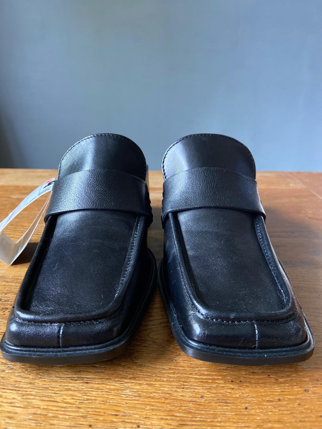 Pair of black leather shoes with a square toe and a strap across the
instep, presented on a wooden table against a grey background.