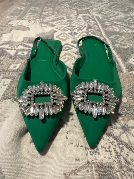 Green pointed-toe shoes with a sparkling rhinestone buckle on a geometric design rug in neutral tones.