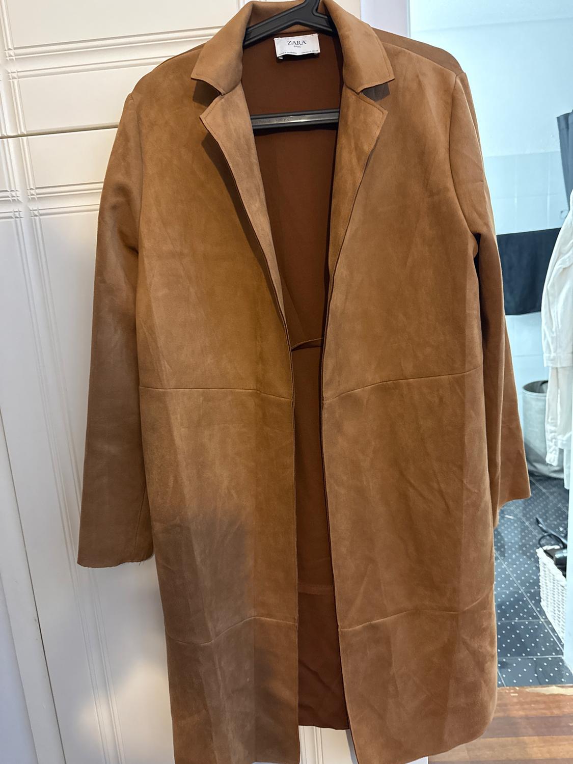 Brown coat hanging on a hanger, with a visible Zara label. White wall background and part of a bathroom.