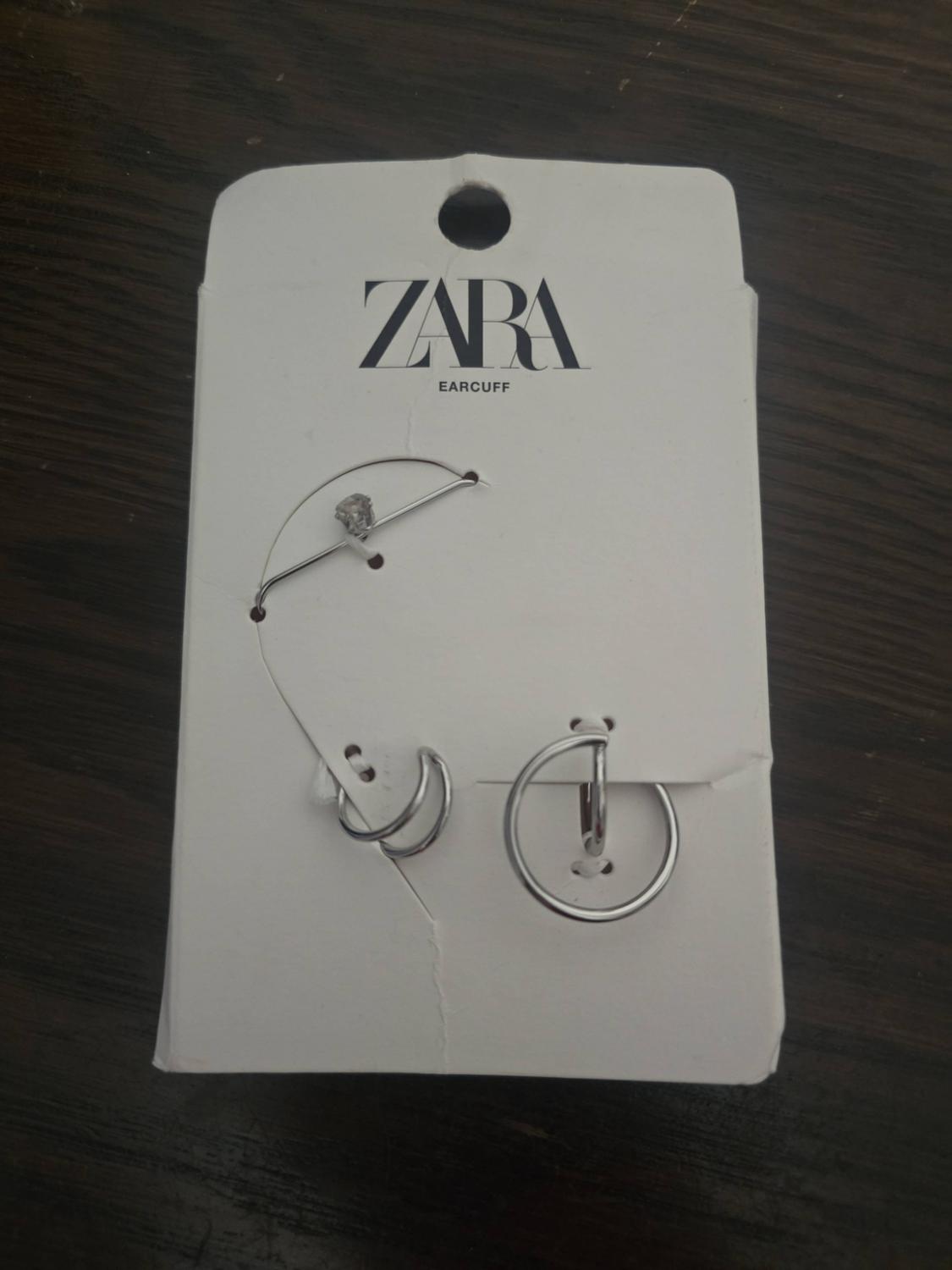 Silver-coloured earrings on a white label against a dark wooden background. Visible text: ZARA EARCUFF.
