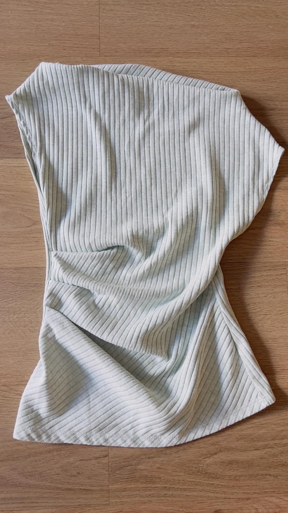 Light-colored ribbed sleeveless top with an asymmetrical design and side draping, on a wooden background.