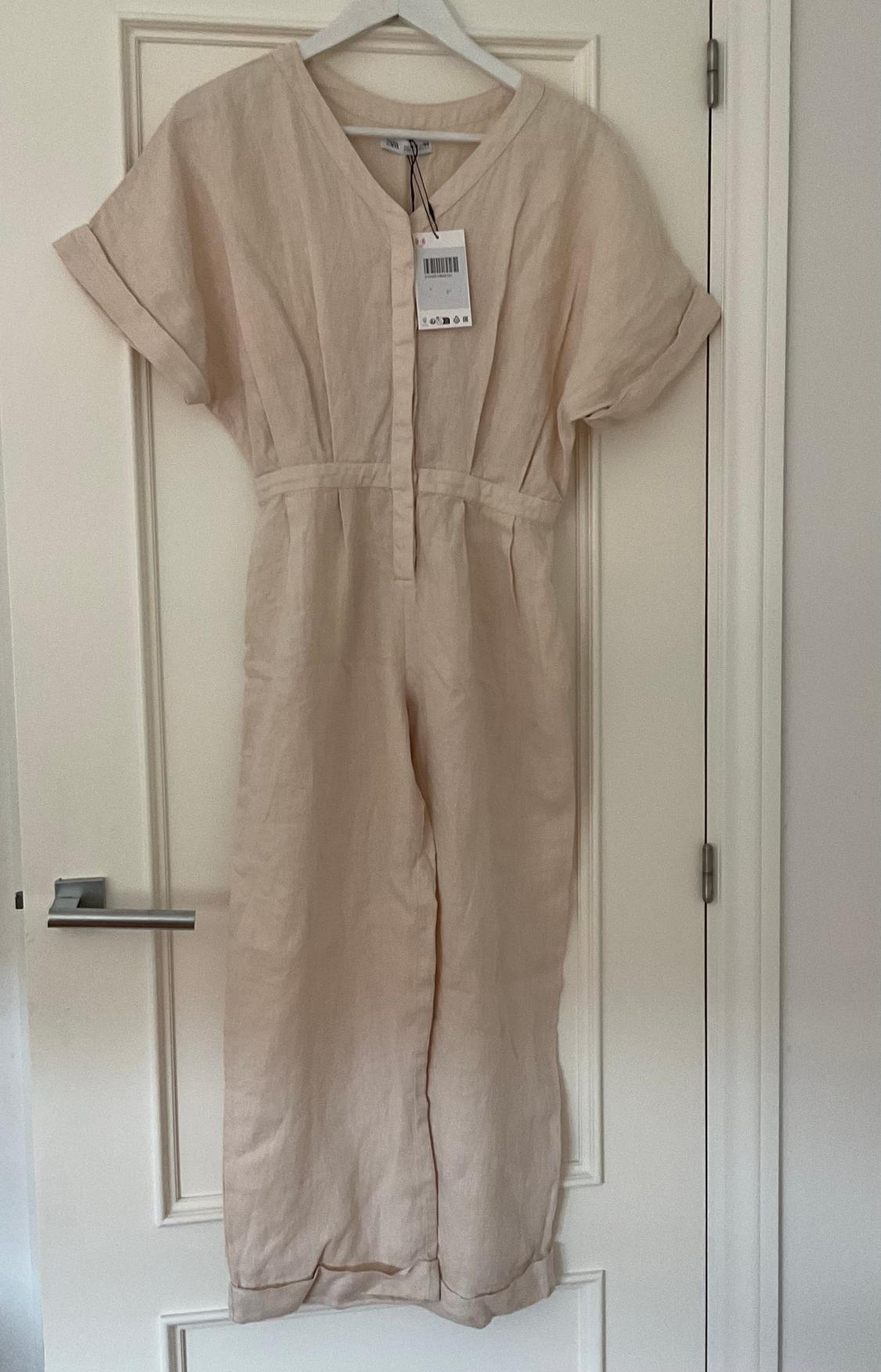 Beige linen jumpsuit hanging on a white door, with visible neck tag. Short sleeves and front fastening.