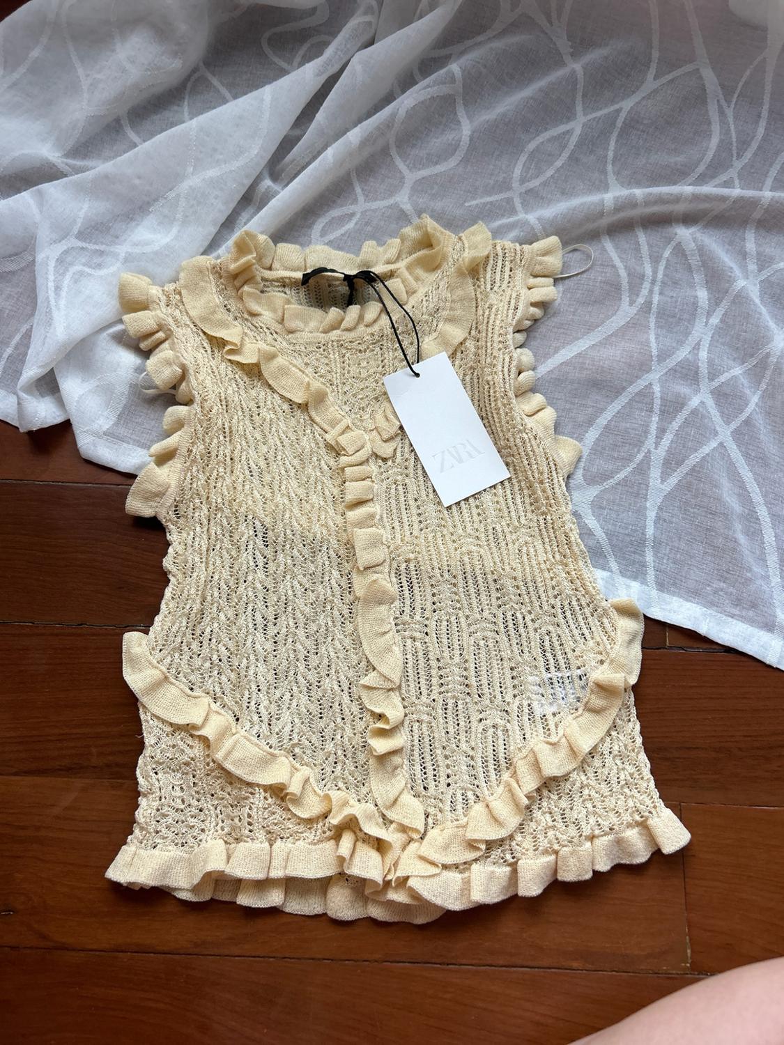 Pale yellow open-knit top with frills and a ZARA label, on a wooden floor and white fabric.