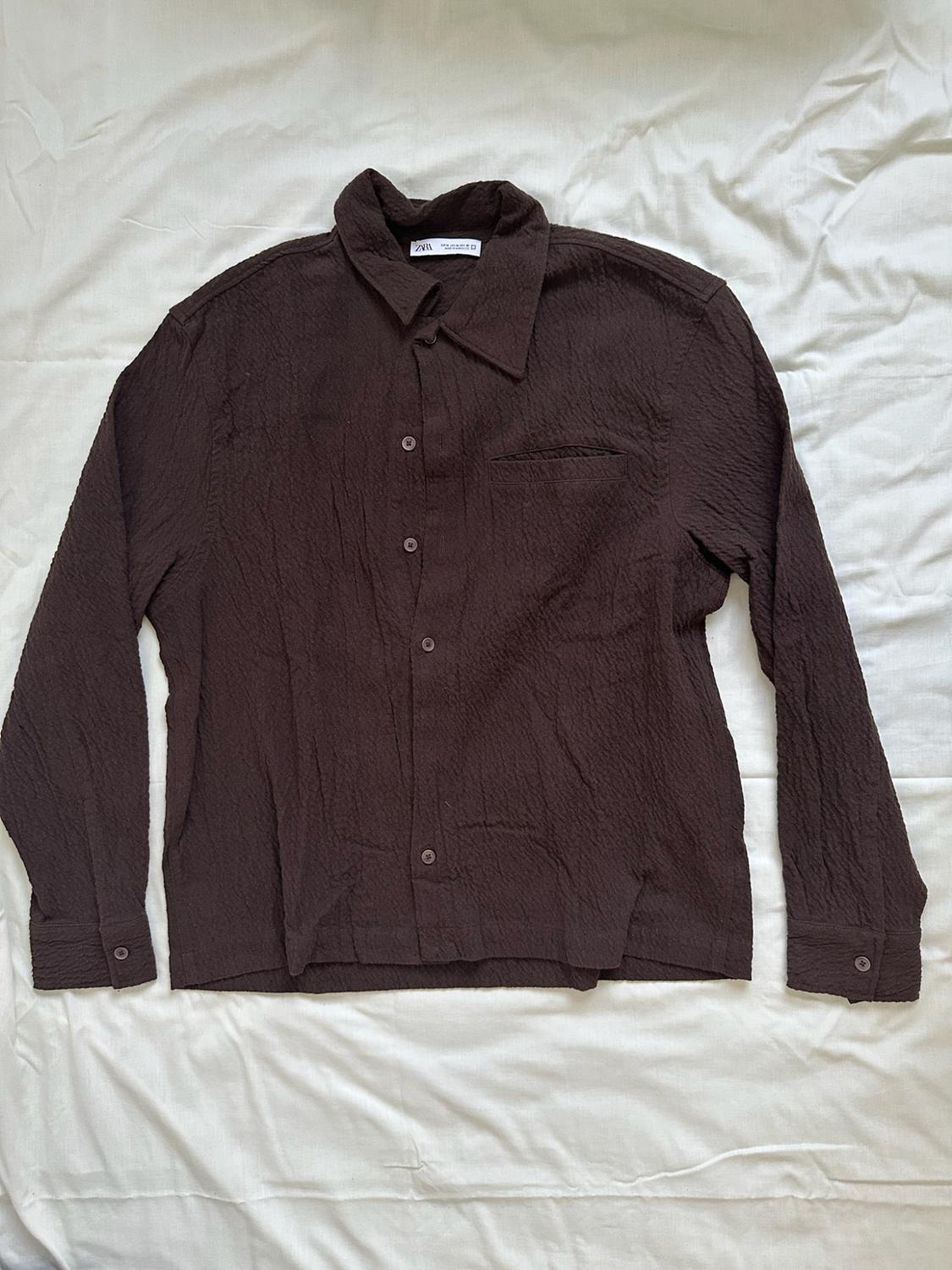 Dark brown long-sleeved shirt with a crinkled texture, laid out on a white fabric. ZARA label visible on the collar.