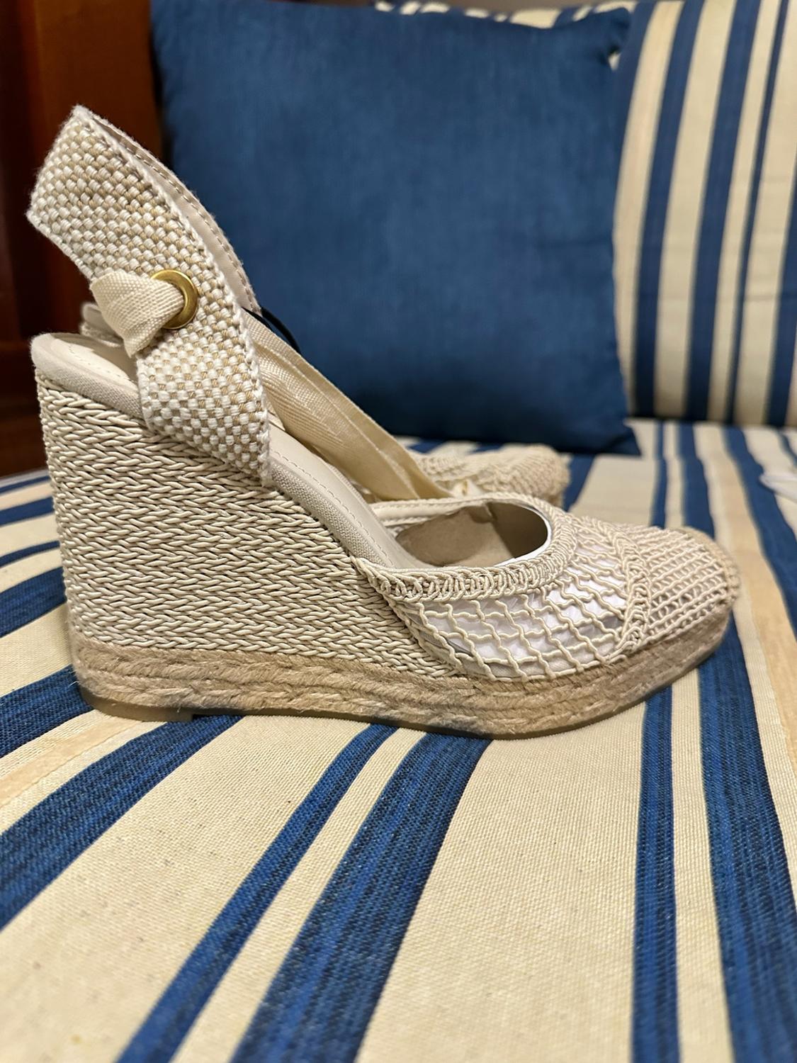 Beige wedge espadrille with woven fabric and mesh detailing, resting on a blue-and-white striped cushion.