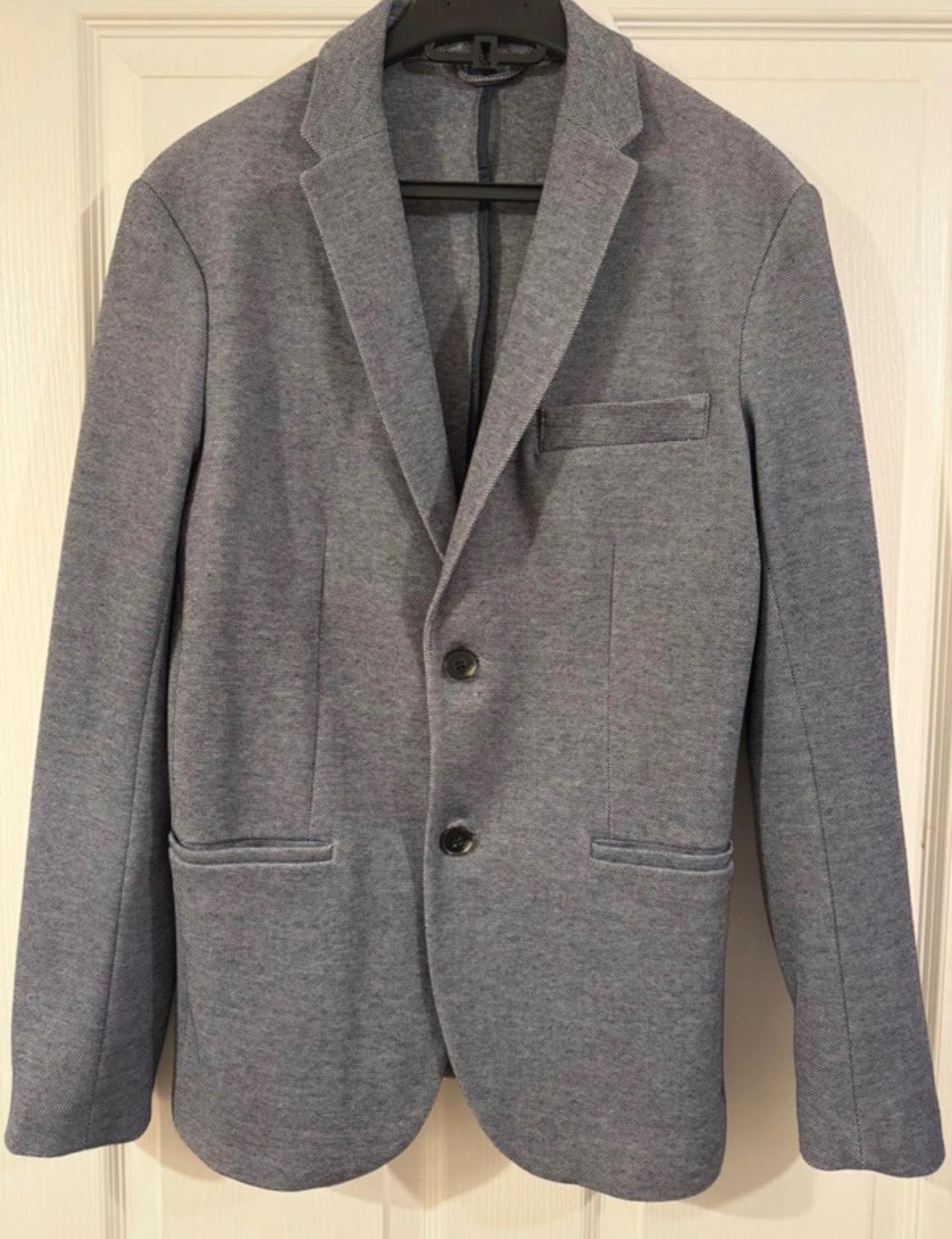 Grey, two-button jacket hanging on a black hanger over a white door. Front pockets and classic lapel.