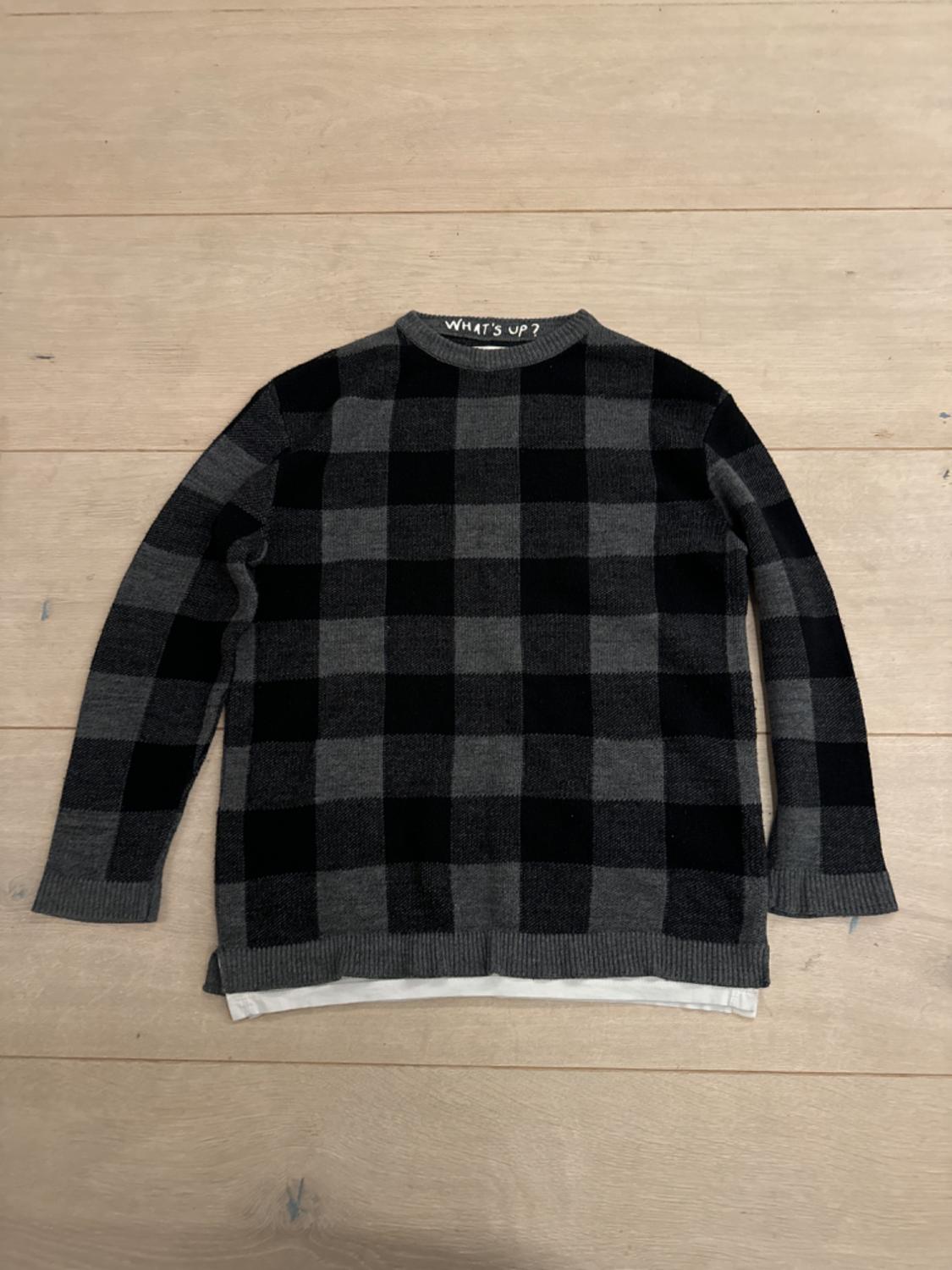 Black and grey checked jumper on a light wooden background. Text "WHAT'S UP?" on the collar.