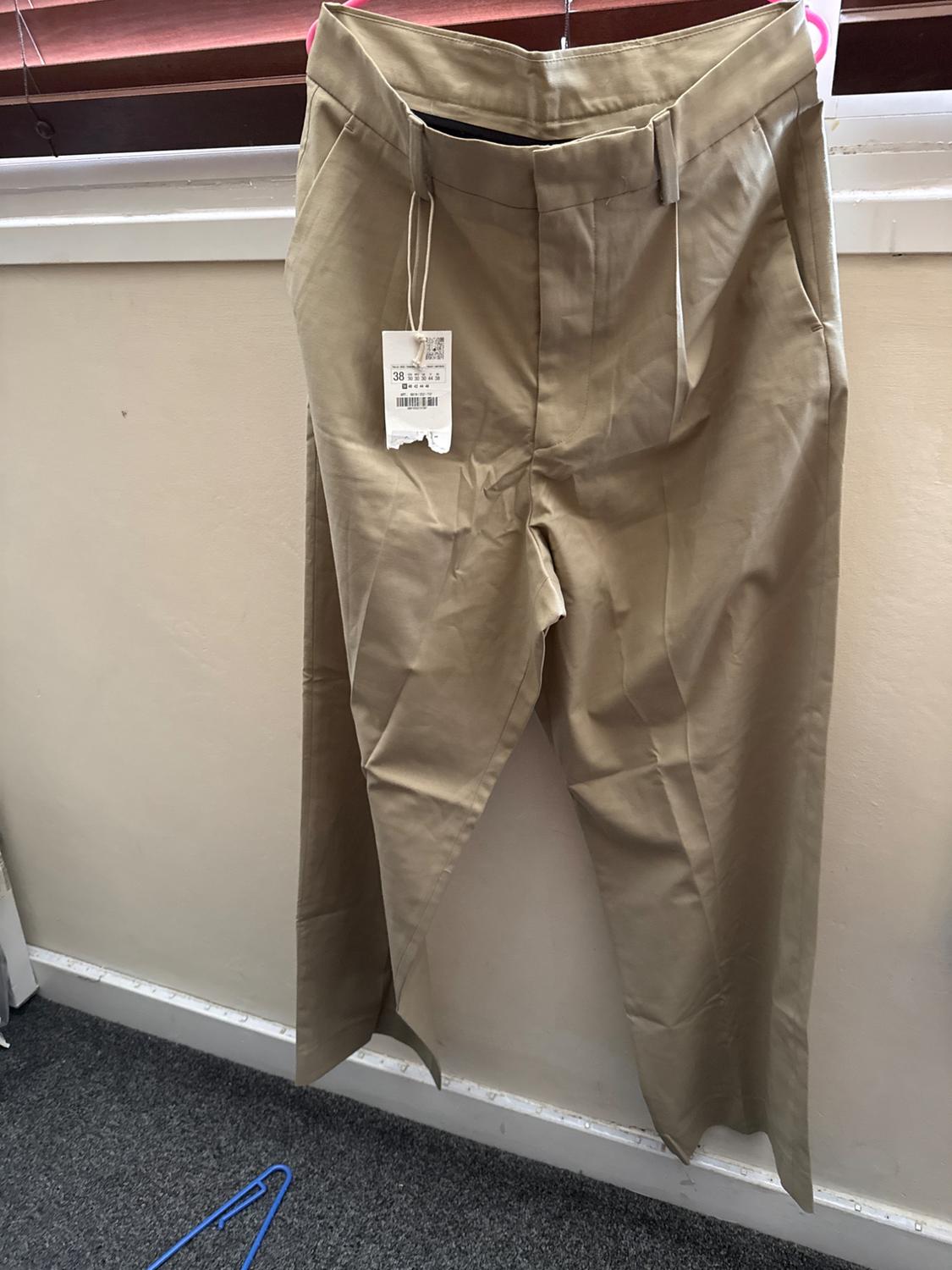 Beige trousers hanging on a pink hanger against a light wall. Visible label with the text "38". Grey floor. Relaxed indoor setting.