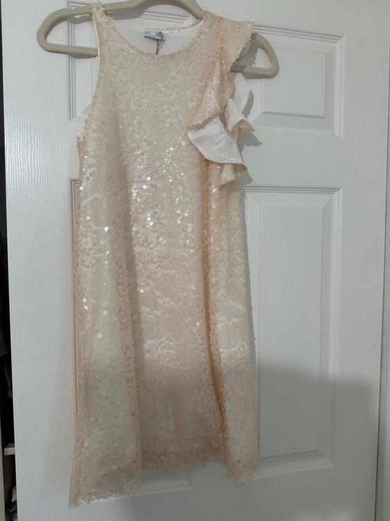 Cream-colored sleeveless dress with sequins and ruffles on the shoulder, hanging on a hanger over a white door.