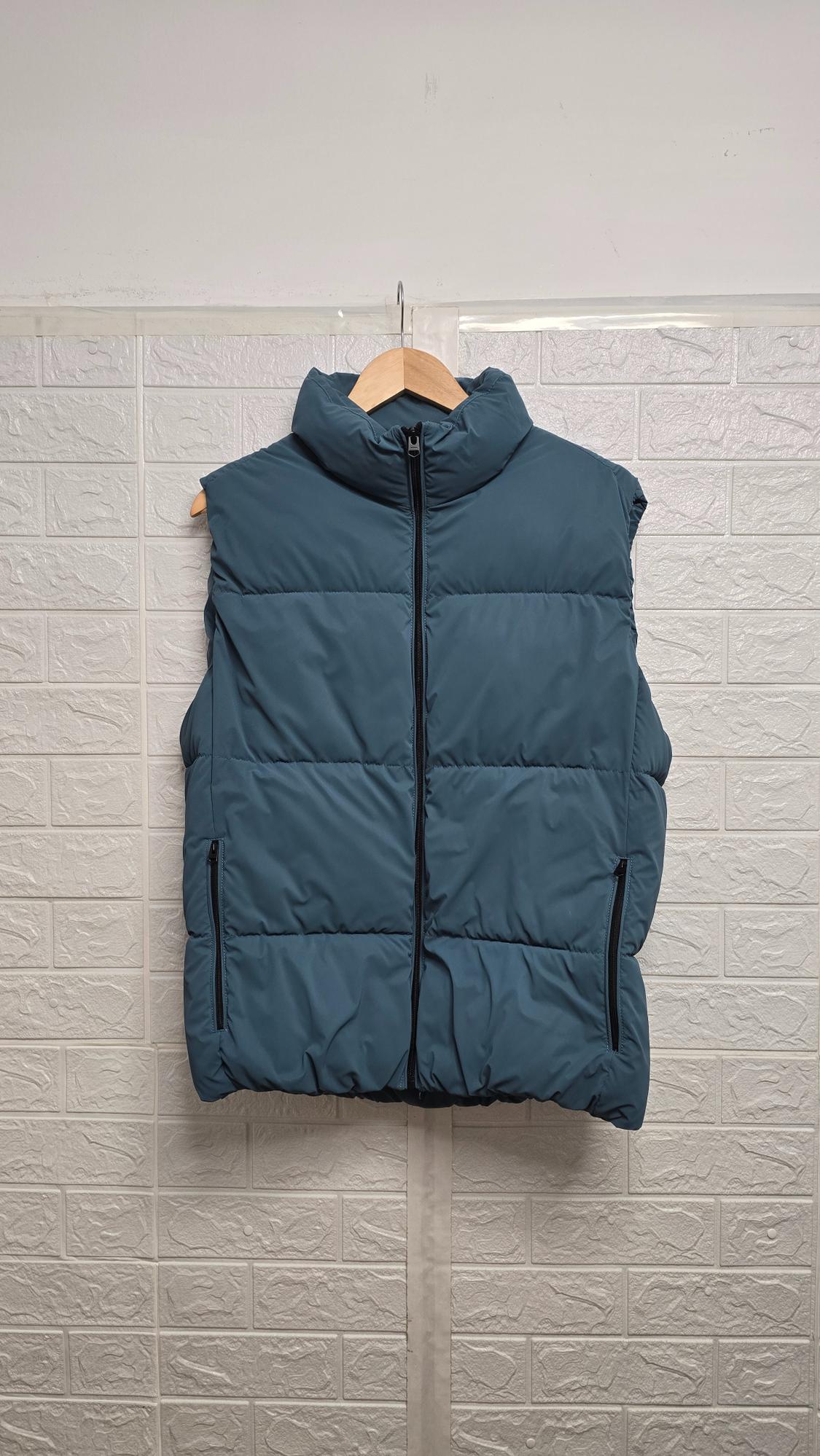 Blue padded gilet with front zip, hanging on a wooden hanger against a white brick-textured wall background.