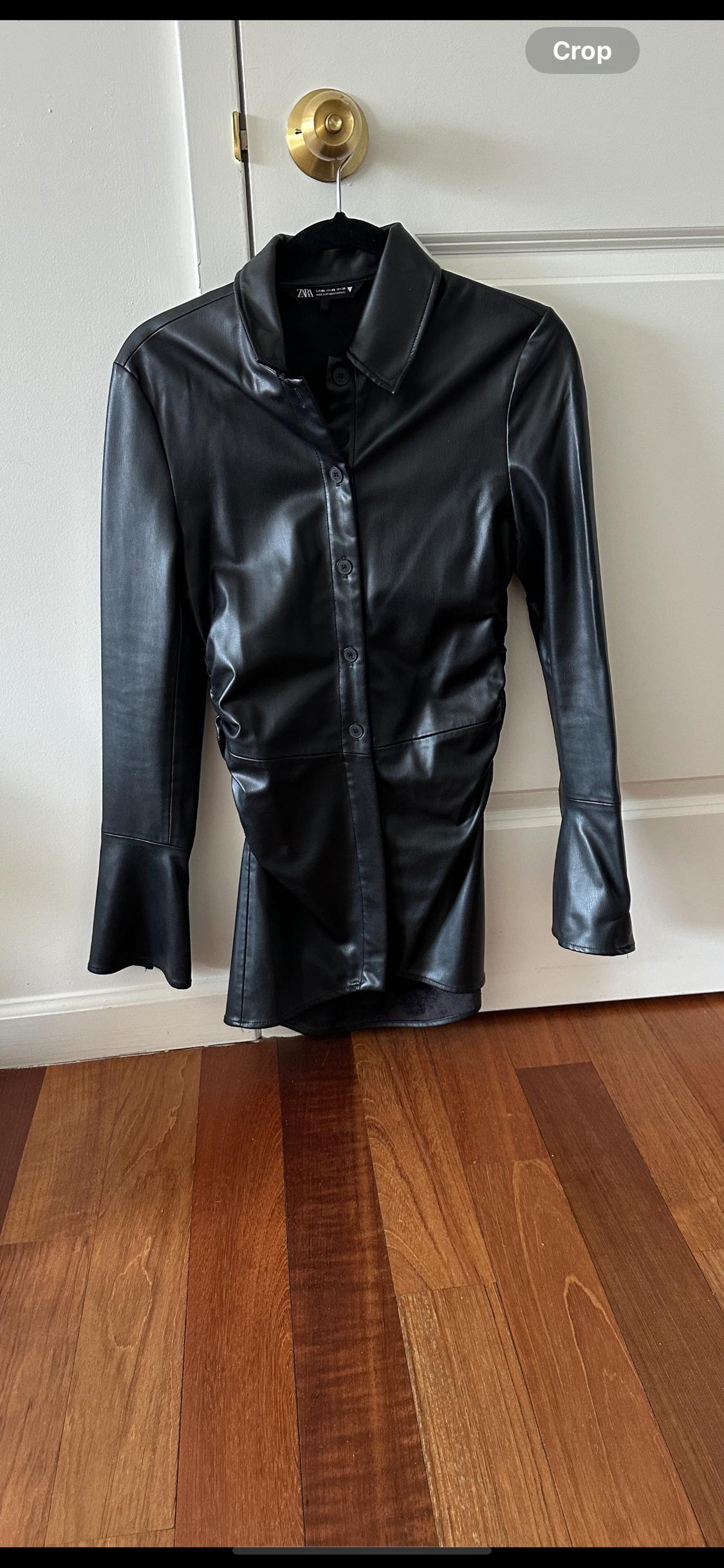 Black leather shirt hanging on a white door with a golden doorknob. The floor is brown wood.