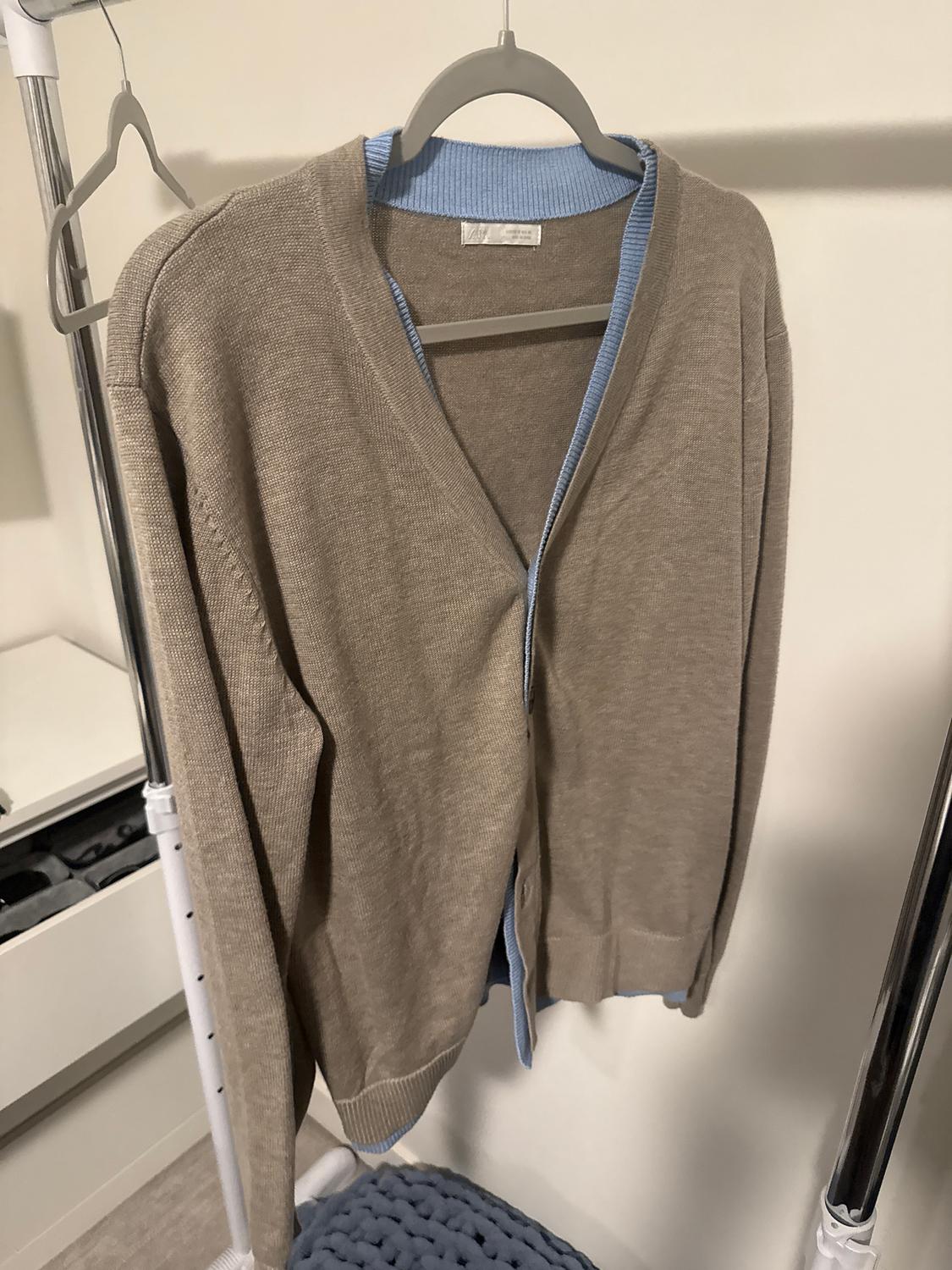 Beige cardigan with light blue trim hanging on a grey hanger, on a clothes rail in a minimalist indoor setting.