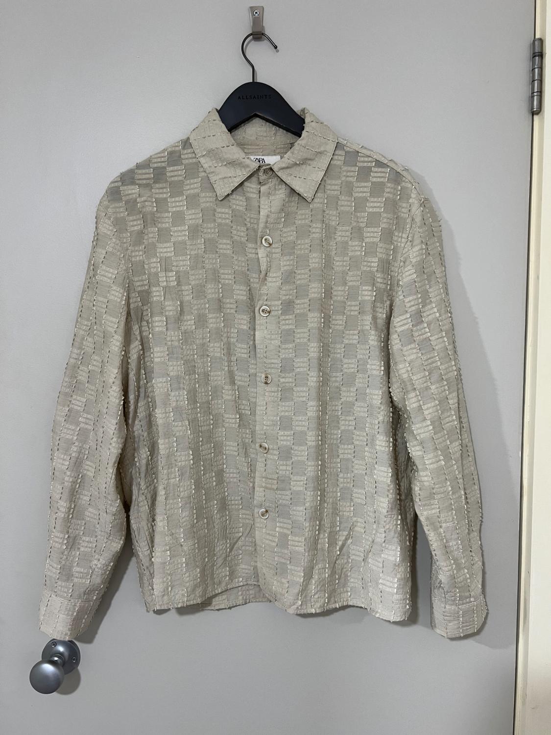 Textured beige shirt hanging on a black AllSaints branded hanger, over a light grey door.