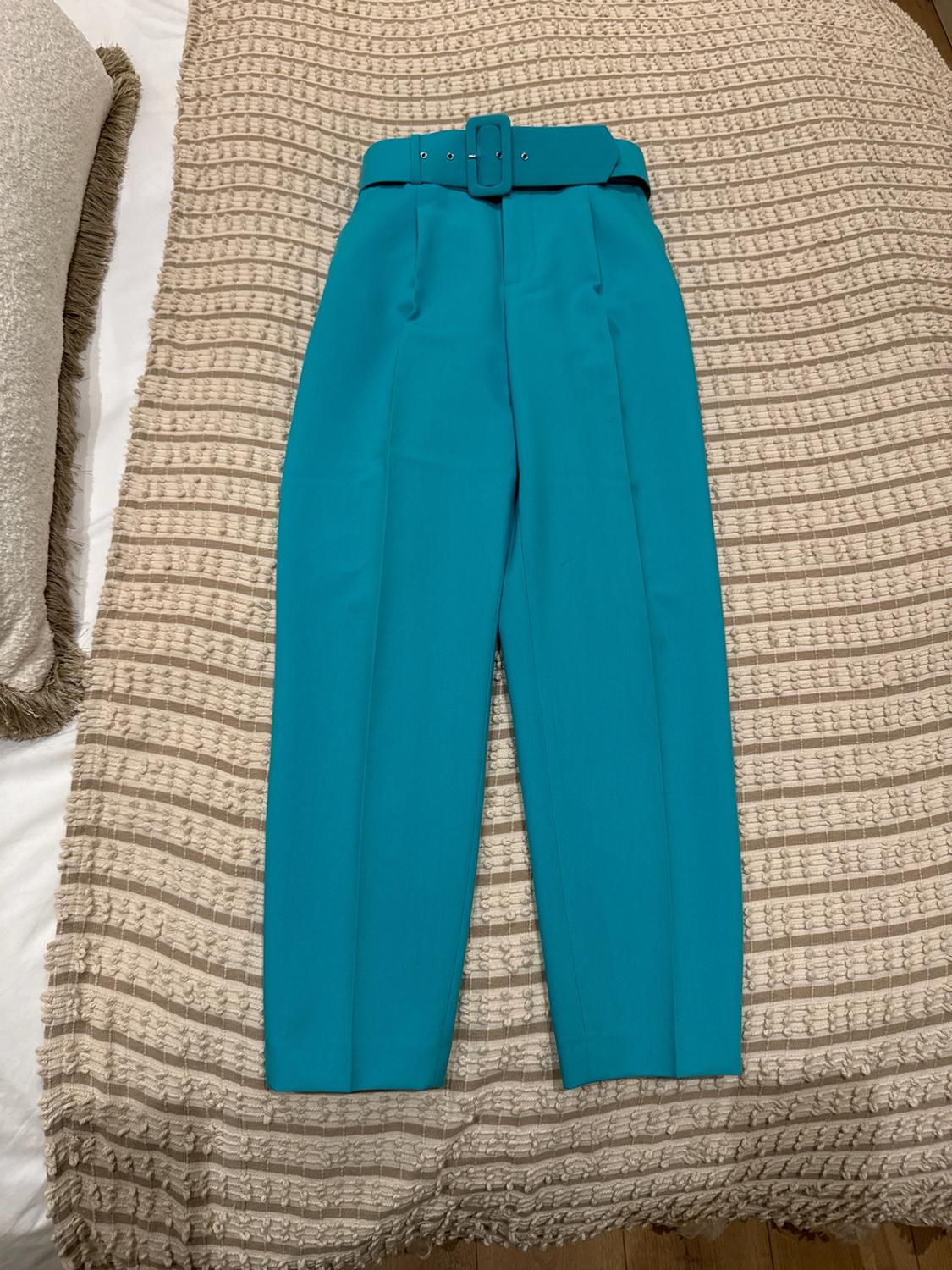 Turquoise pleated trousers with a matching belt, laid out flat on a beige striped blanket on a bed. Relaxed atmosphere.