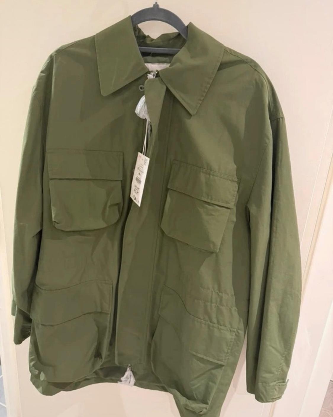 Olive green jacket hanging on a hanger against a light background. Design with a collar, front zip fastening and four large flap pockets.