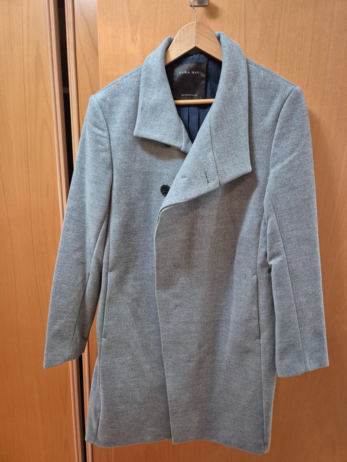 Men's grey coat hanging on a wooden hanger, with a wide lapel and two visible buttons, against a light wood background.