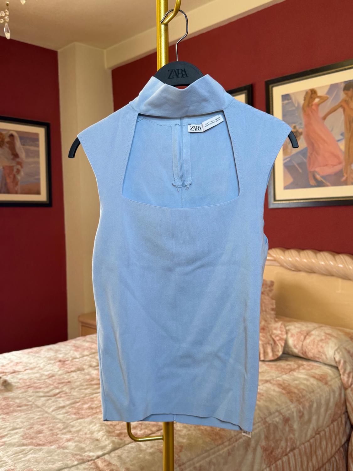 Blue sleeveless top hanging on a black hanger with a "ZARA" tag. Red wall background and bed with a patterned bedspread.