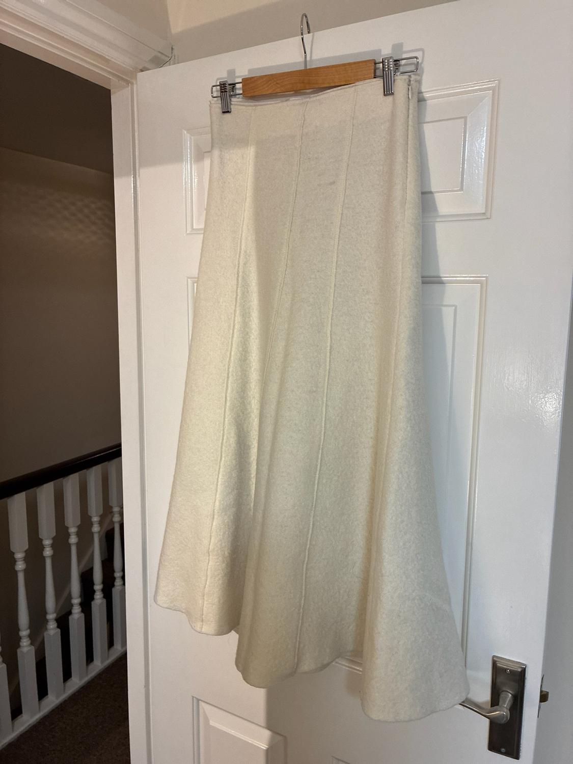 Cream-coloured long skirt with vertical stitching, hanging from a wooden hanger on a white door. A staircase is visible in the background.