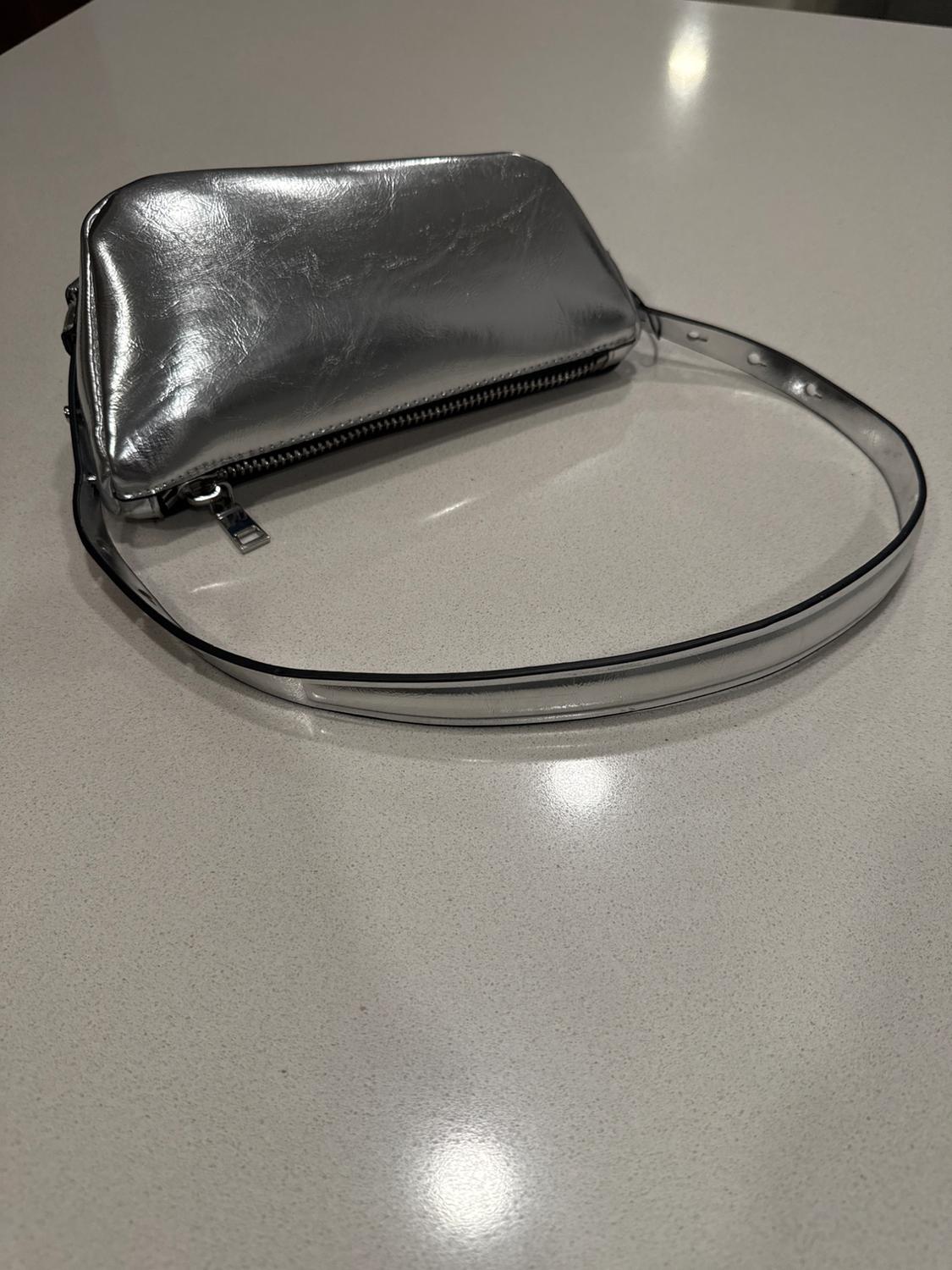 Silver handbag with zip and thin strap on a light surface. Shiny reflections on the material.