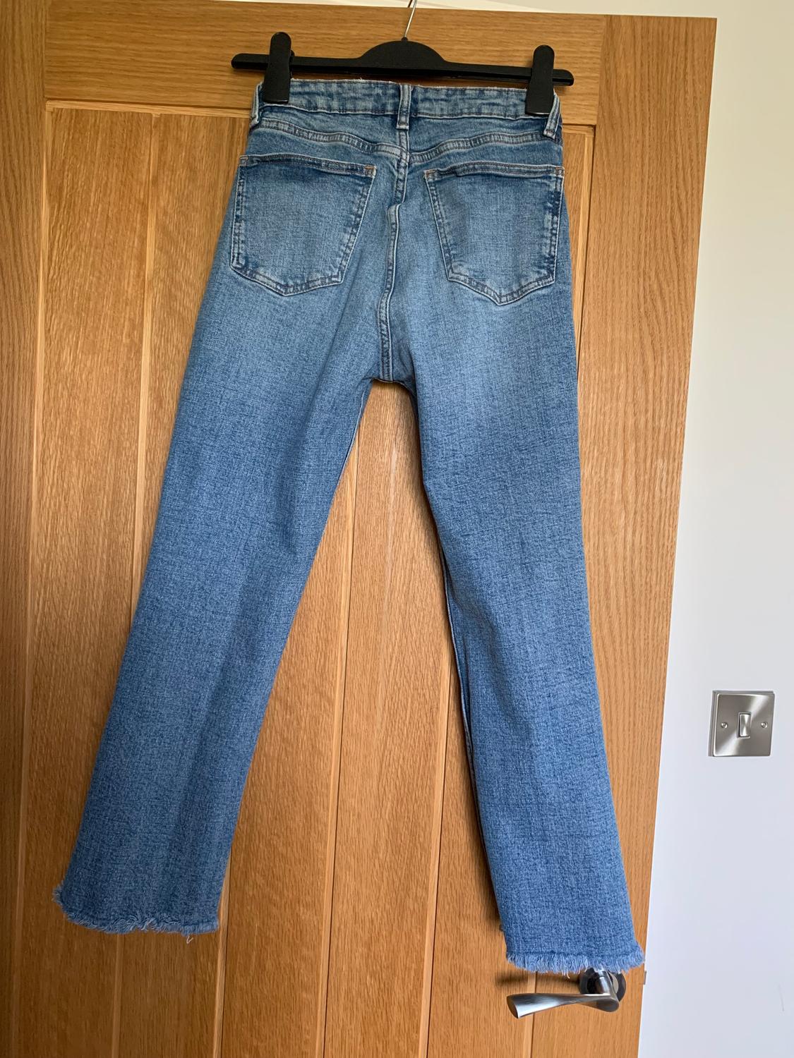 Blue jeans hanging on a wooden door, seen from behind. They have a straight-cut design and frayed hem.