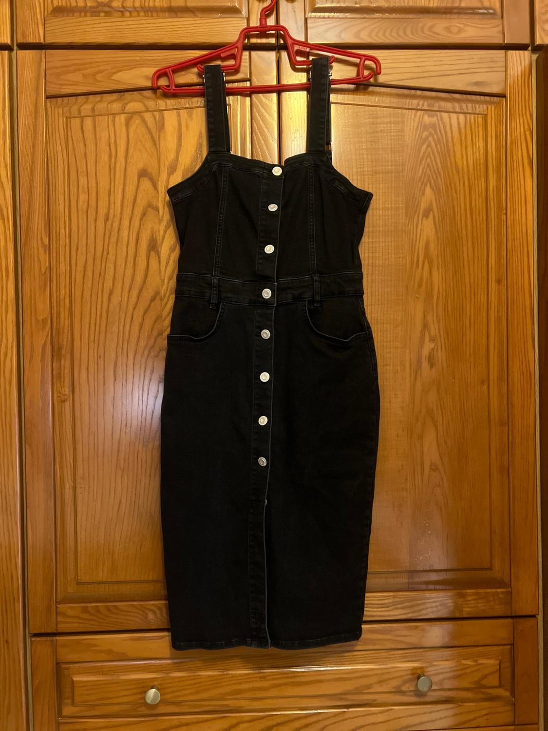 Black pinafore dress with front buttons and pockets, hanging on a red hanger against a wooden wardrobe. Casual and relaxed vibe.