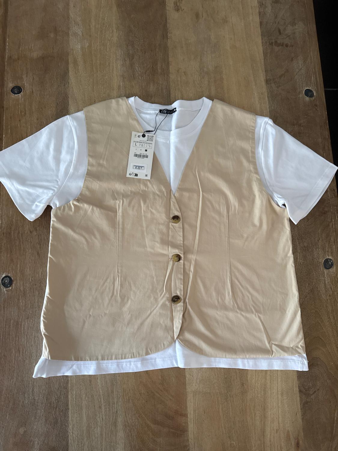 Beige button-down waistcoat over a white t-shirt, placed on a wooden table. Visible hanging tag with product details.