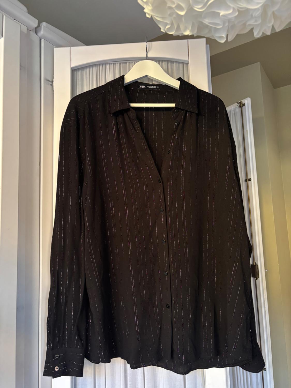 Long-sleeved black shirt with thin, sparkly lines, hanging on a white hanger in front of a white door with pleated curtains.