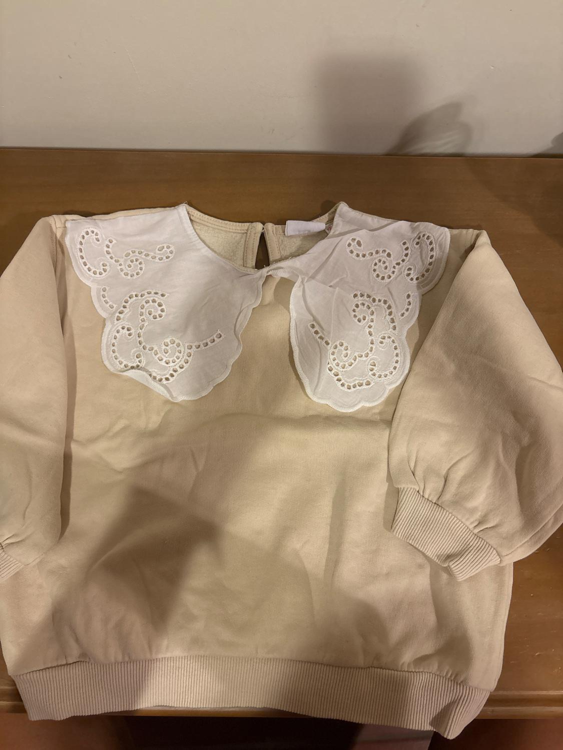 Beige sweatshirt with embroidered white collar, on a wooden table. Long sleeves and elasticated cuffs.