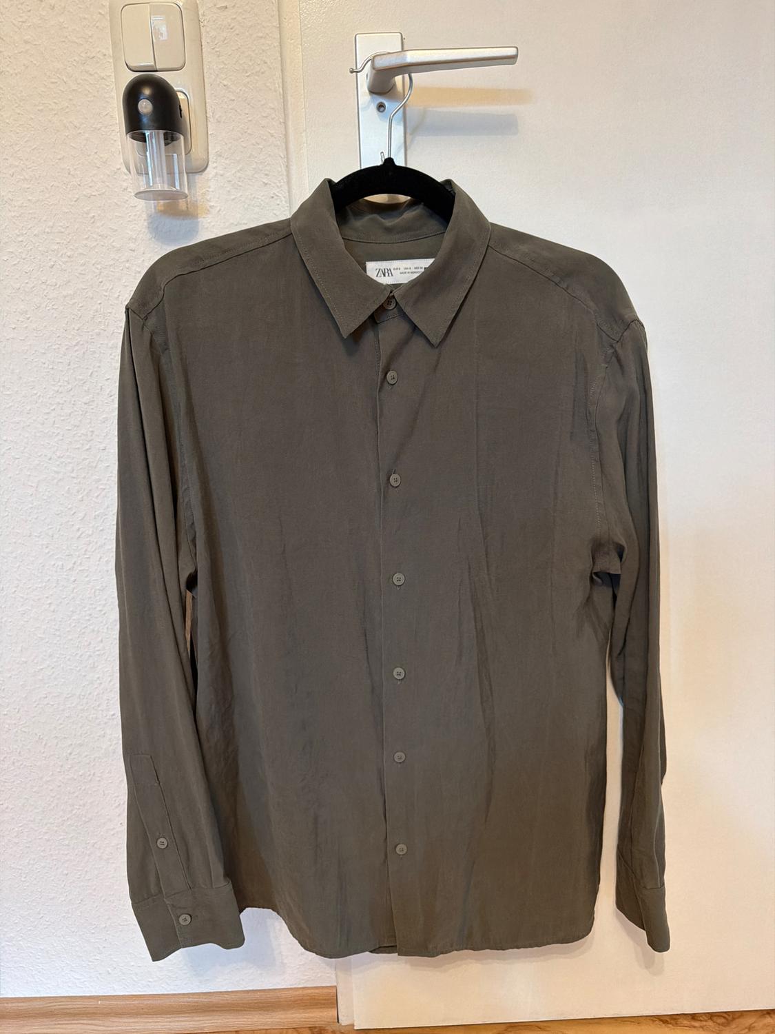 Khaki long-sleeved shirt hanging on a black hanger over a white door. Label visible on the collar.
