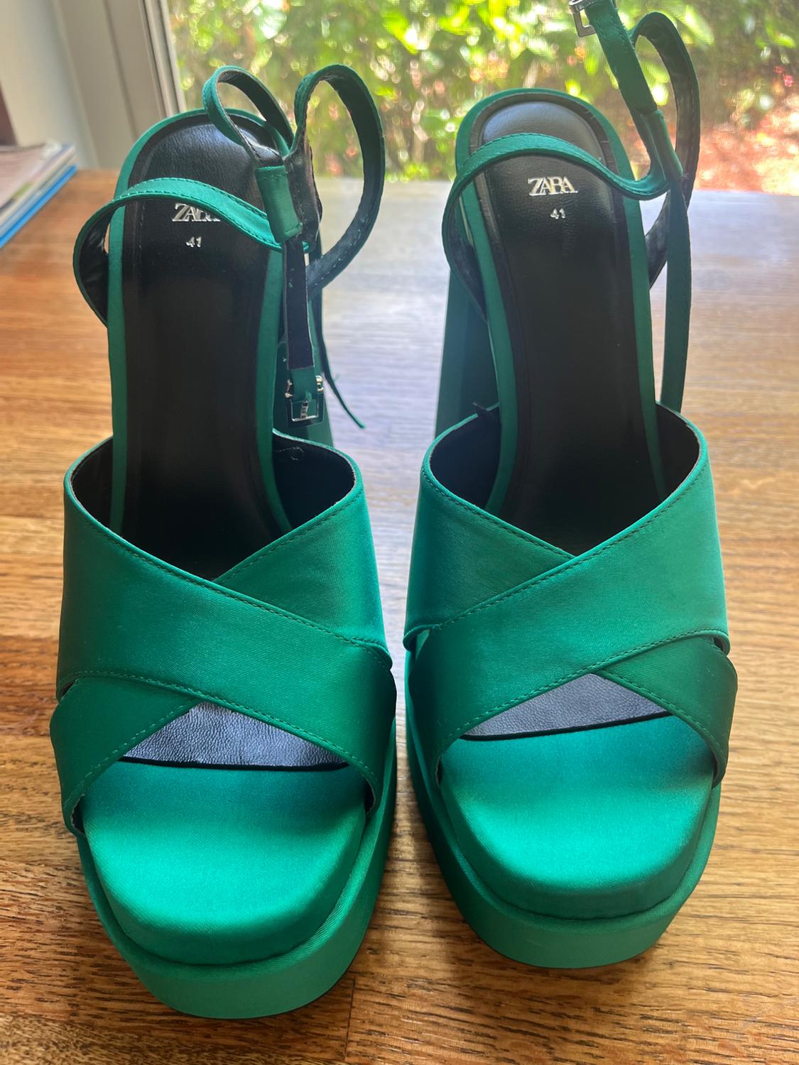 Green heeled and platform sandals with crossed straps on a wooden table in front of a window. Text: ZARA 41.