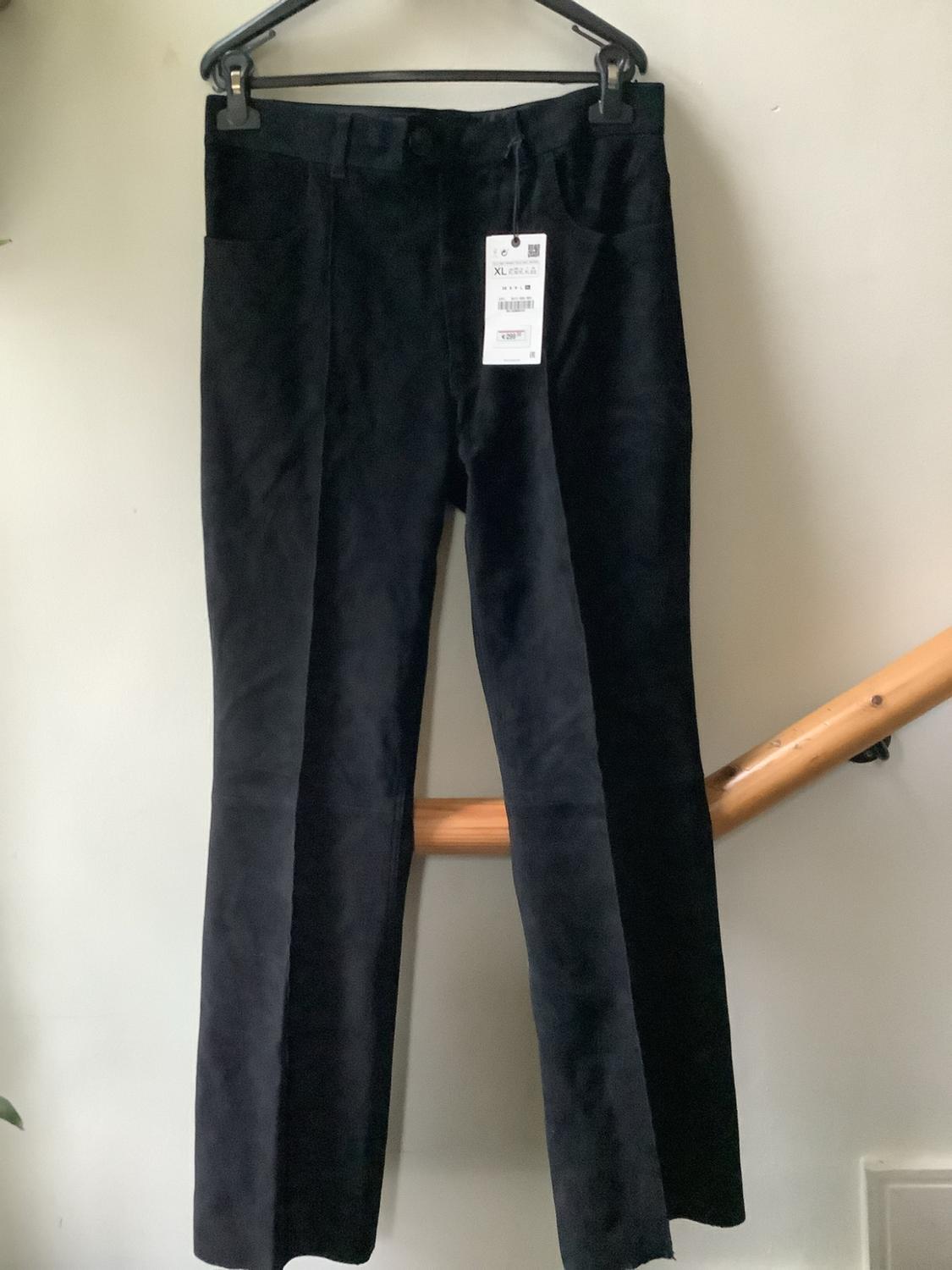Black trousers hanging on a coat hanger against a white wall. They have a label bearing the text “XL”. A wooden handrail can be seen in the background.