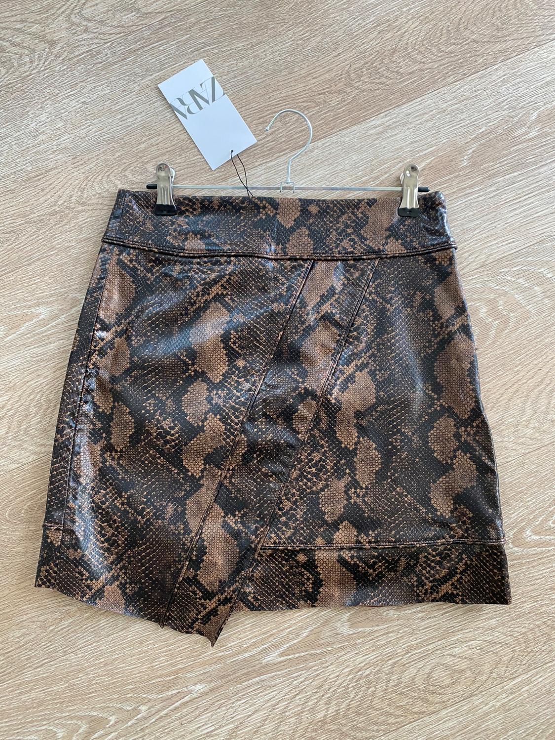 Short skirt with snake print in brown and black tones, hanging on a hanger over a light wood floor. Visible tag.