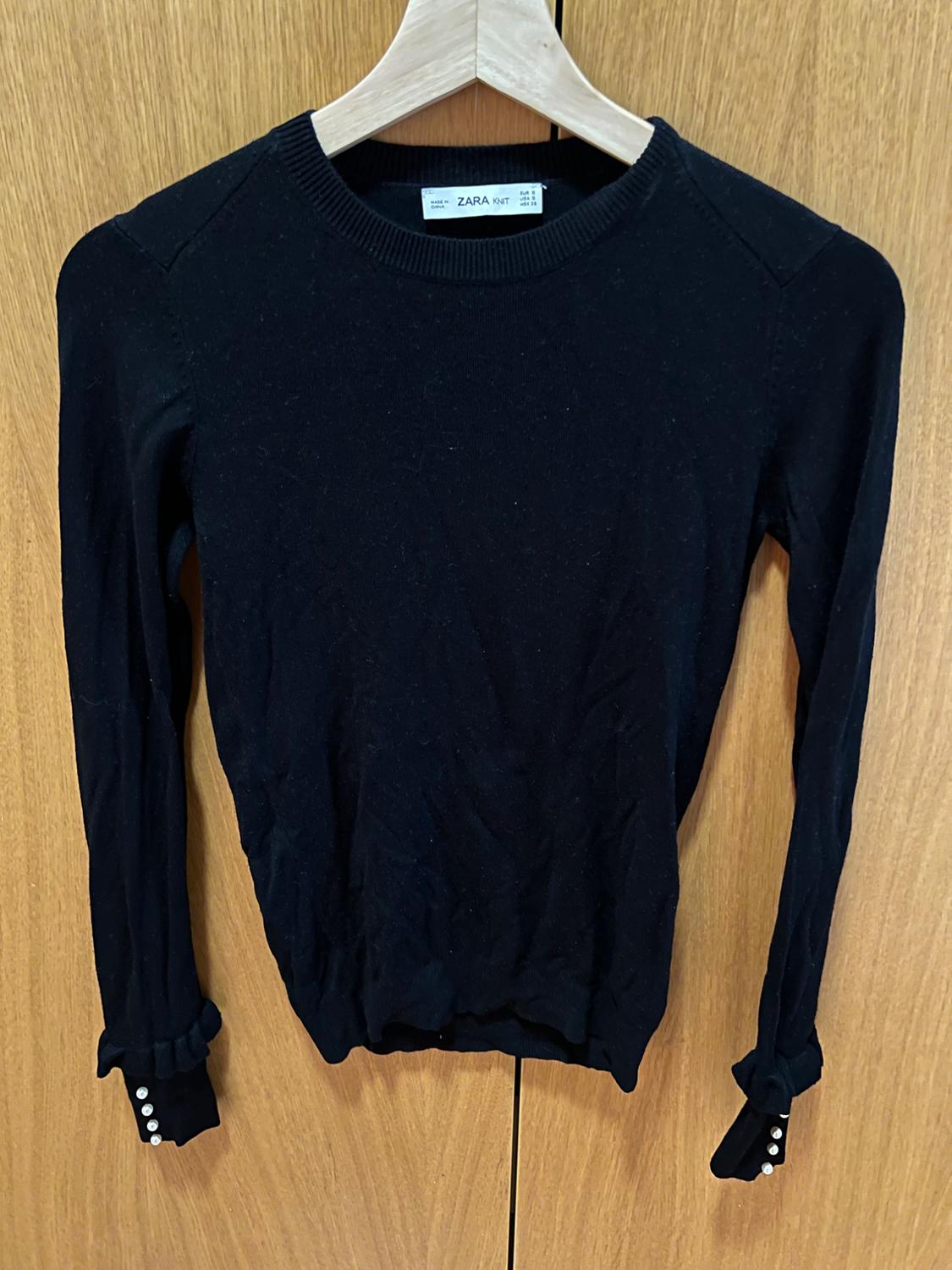 Black jumper hanging on a hanger against a wooden background. Long sleeves, round neck, cuffs with frills and buttons. Text: ZARA KNIT.