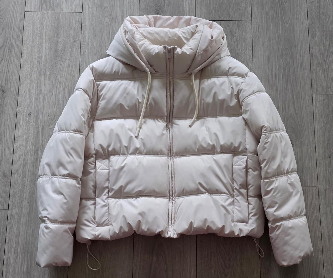 White puffer jacket with hood and drawstrings, on gray wood floor. Horizontal panel design and zipper closure.
