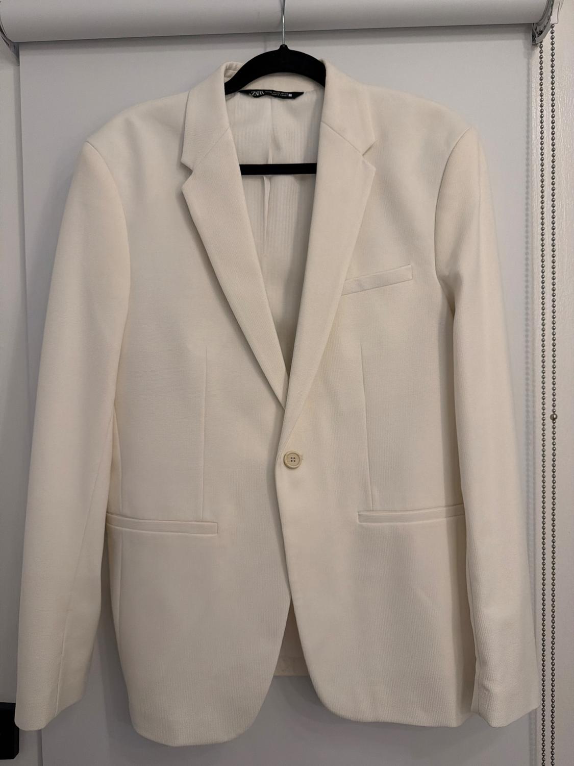 White single-button blazer hanging on a black hanger in front of a white door. Interior label with the text ZARA.