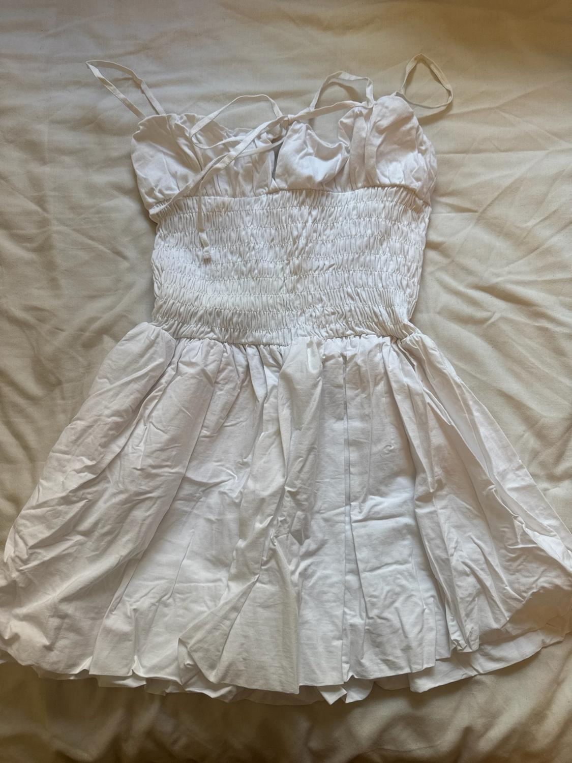 Short white strappy dress with a gathered bodice, laid out on a light, crumpled sheet. Casual and relaxed style.