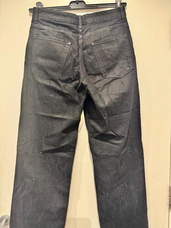 Black trousers, hung and viewed from behind, with a wavy line design and shimmering details throughout the fabric.