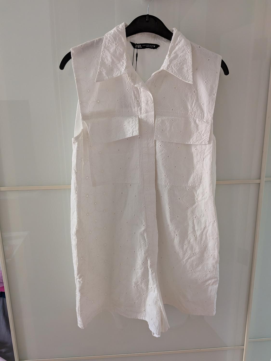 Sleeveless white dress with floral embroidery, classic collar, and chest pockets, hanging on a black hanger.