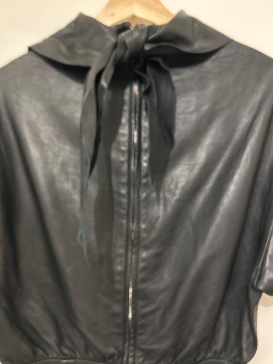 Black leather jacket hanging on a wooden hanger, with a central zip and a bow at the neck.