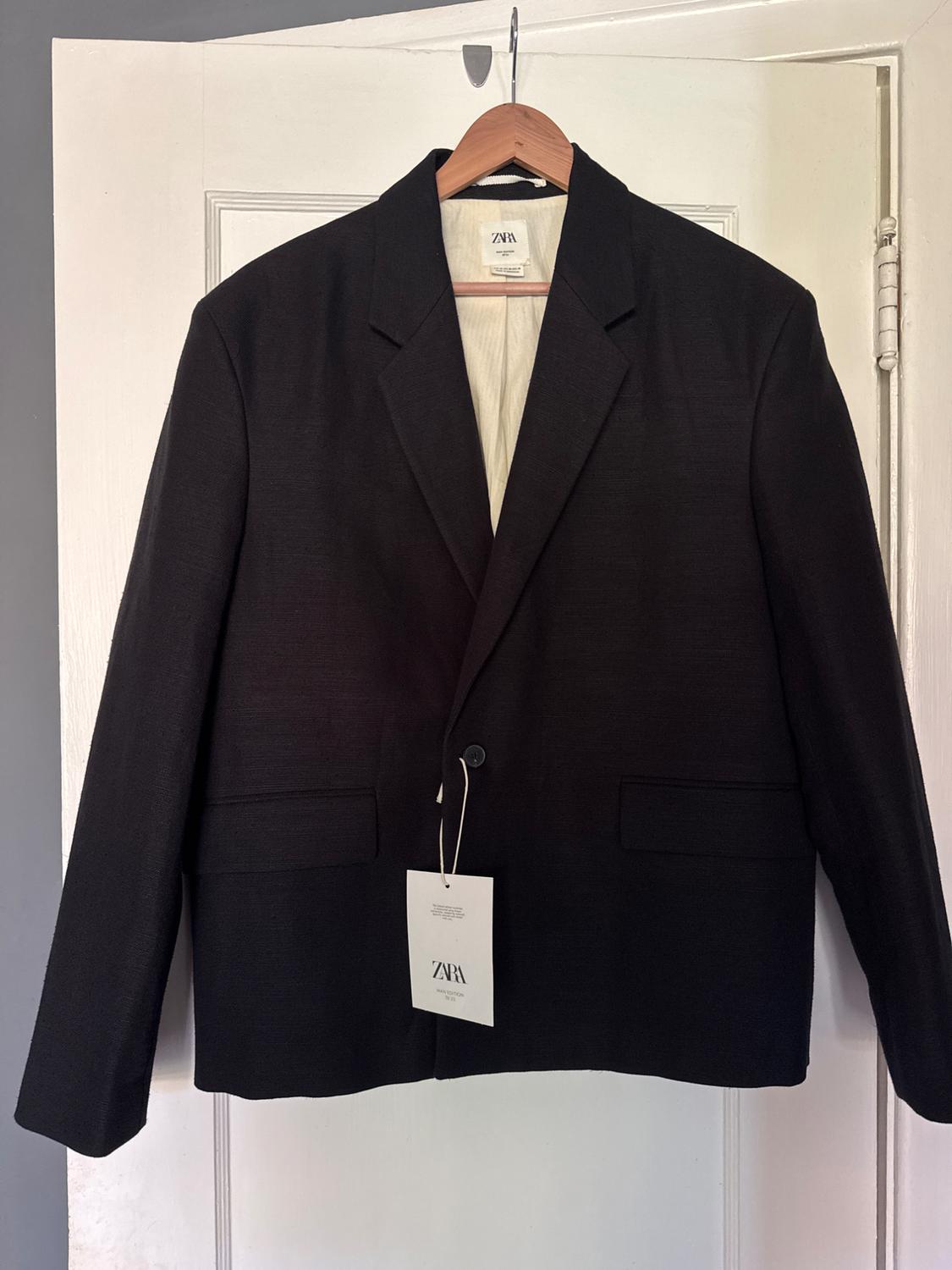 Black suit jacket hanging on a wooden hanger. Visible label with the text "ZARA". White door in the background.