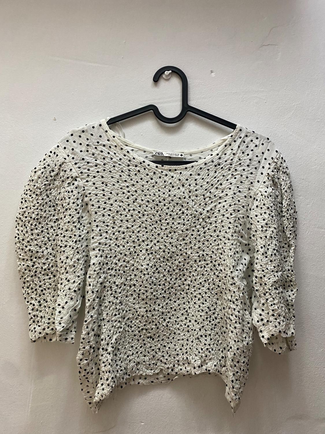 White, long-sleeved blouse with a black polka dot print, hanging on a black coat hanger against a white wall.