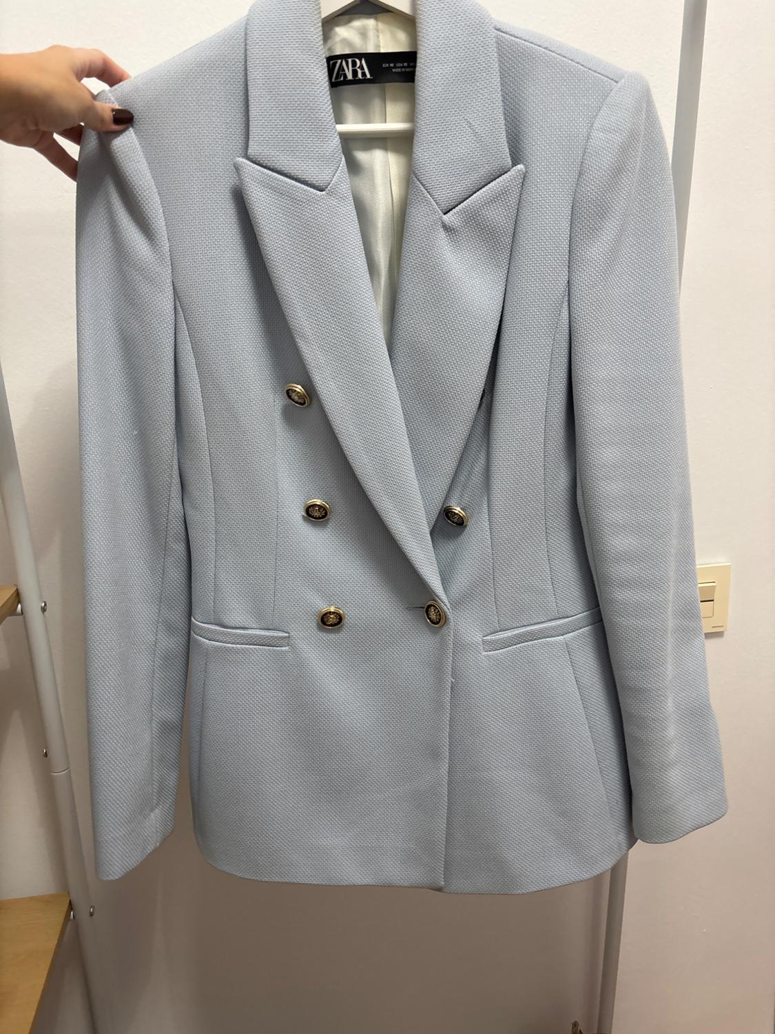 Light blue double-breasted jacket with gold buttons, hanging on a hanger. Visible label: ZARA. Hand holding the jacket.