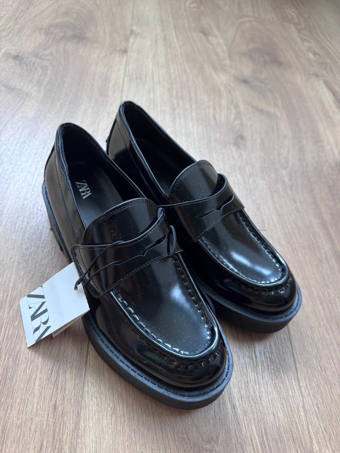 Shiny black loafers with thick soles on a wooden floor. The text ZARA can be read inside them and on a white label.