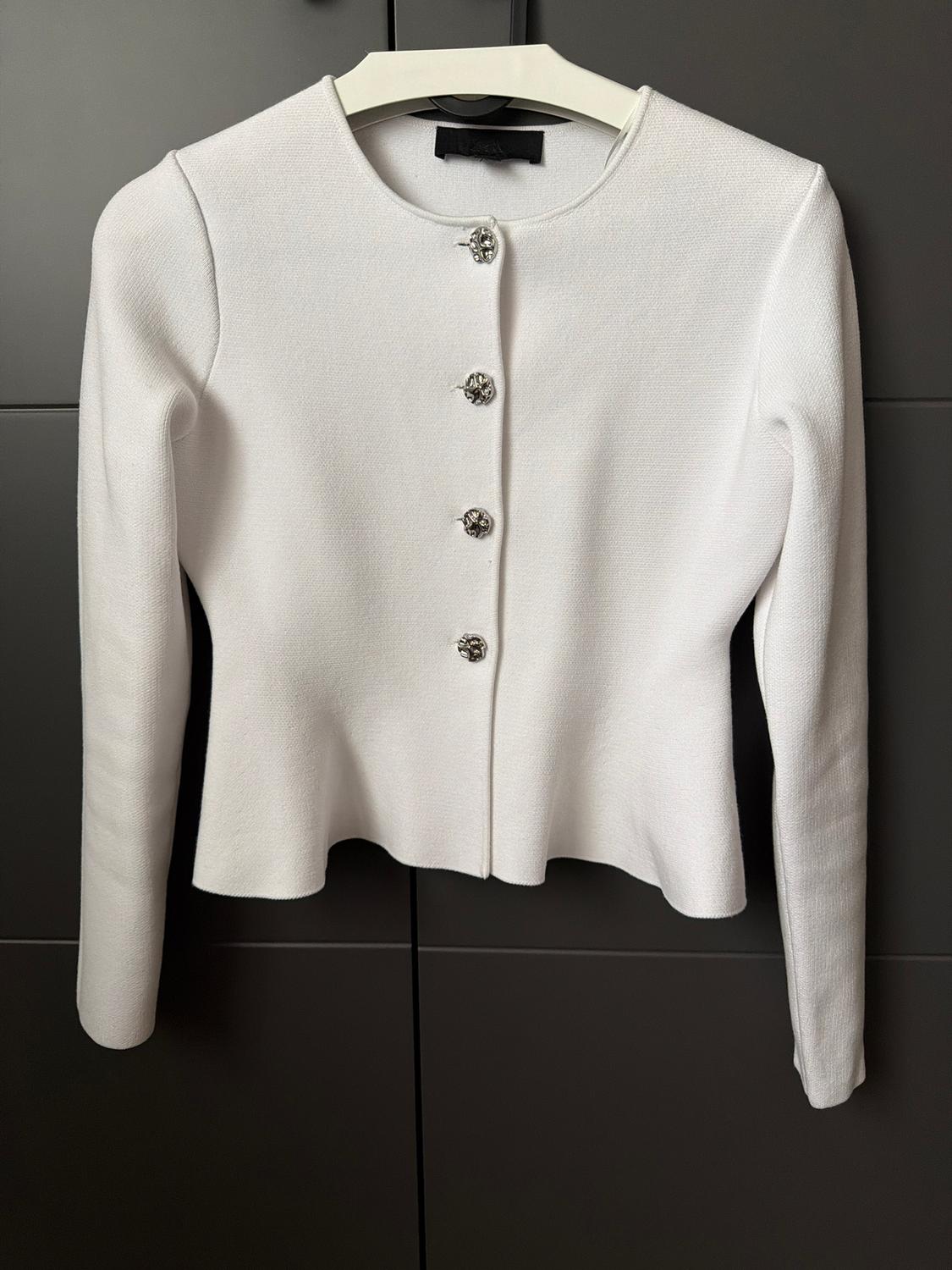 White, long-sleeved jacket with decorative silver buttons, hanging on a hanger against a dark background.