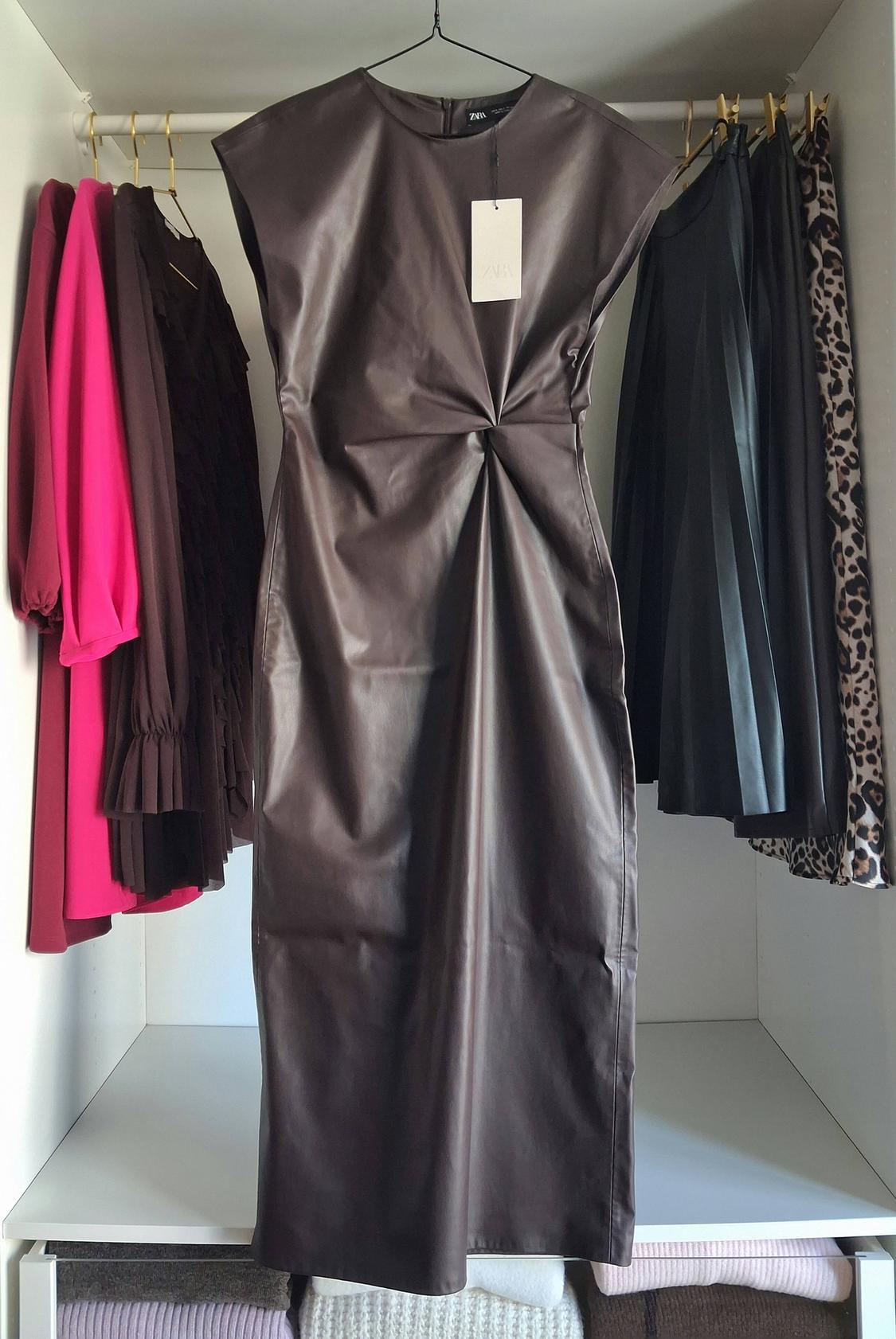 Brown sleeveless dress hanging in a wardrobe next to pink, brown, and animal print garments. Visible tag.
