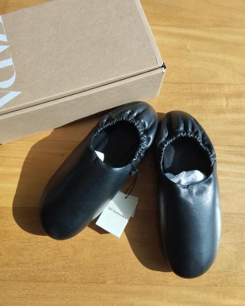 Black leather shoes with elasticated sides next to a cardboard box on a wooden surface. Label visible on one shoe.