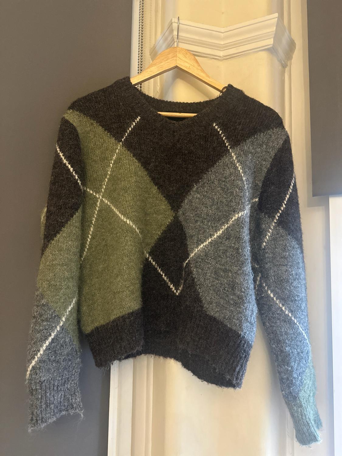 Knitted jumper with diamond patterns in dark grey, green and light blue, hanging on a wooden hanger against a grey wall.