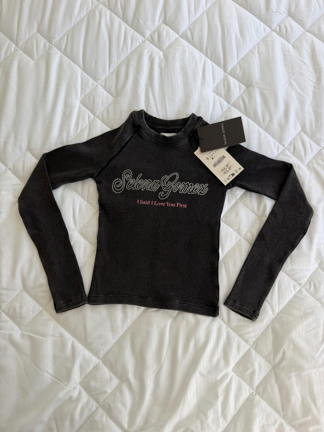 Black long-sleeved jumper on white quilt, with "Selena Gomez I Said I Love You First" text on the front and hang tags.