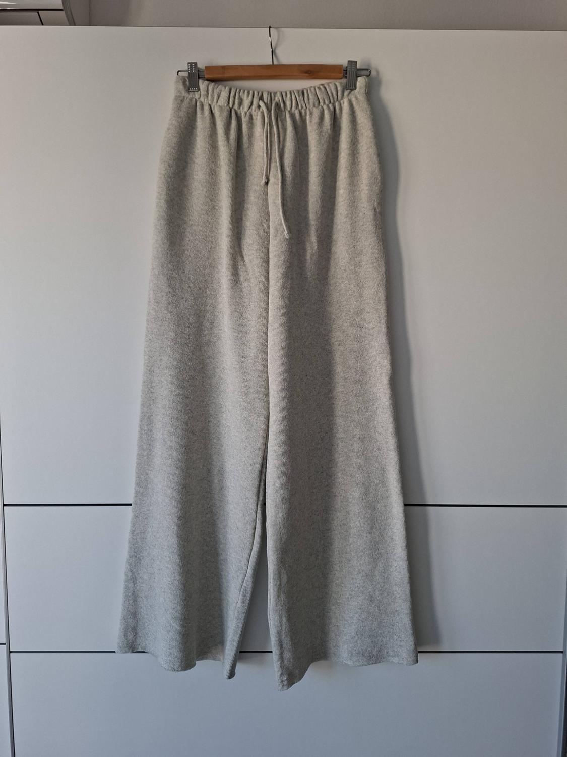 Light grey knitted trousers with wide legs and an elasticated drawstring waist, hanging on a wooden hanger against a white background.